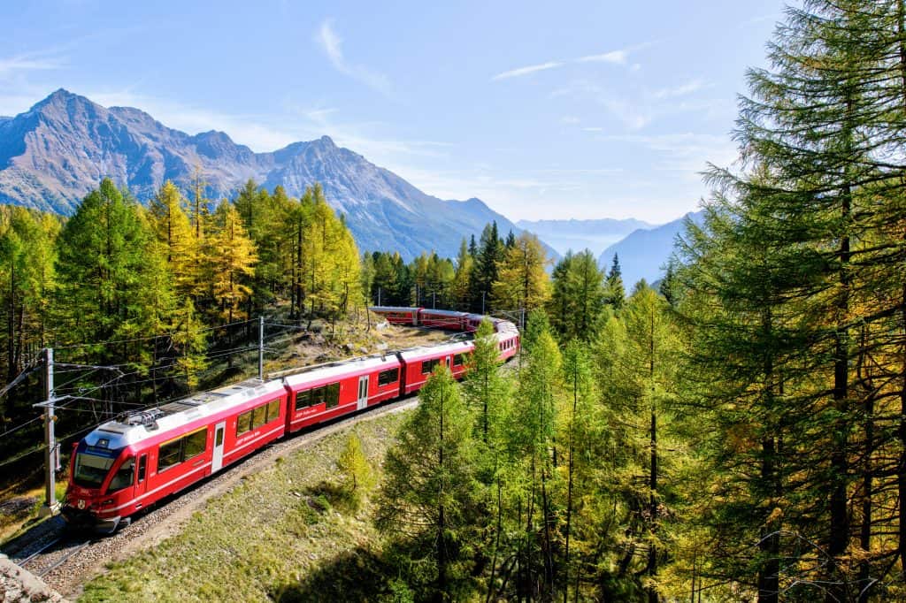 Red train traveling through lush green forest with mountains in the background.